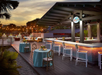 Beachside bar at sunset with tables and chairs on a wooden deck, string lights above, and a calm atmosphere.