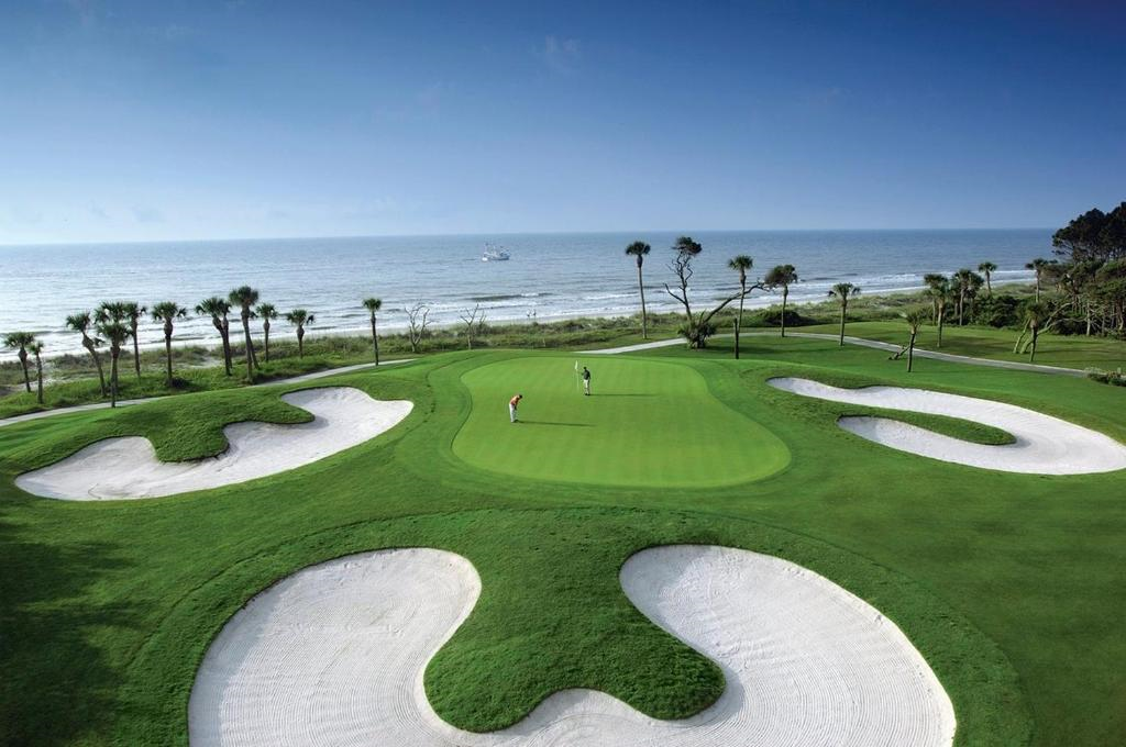 A golf course with sand bunkers overlooks the ocean. Two people are standing on the green, with a ship visible on the horizon under a clear sky.