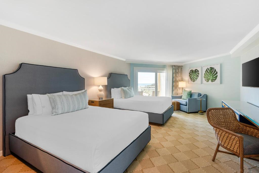 A hotel room with two double beds, a blue armchair, a wicker chair, a wall-mounted TV, and a balcony view. The decor is light and airy with tropical-themed artwork on the wall.