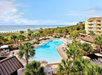 Aerial view of a beachfront resort with a large pool, surrounded by palm trees and sun loungers. Ocean waves and beach umbrellas are visible in the background.