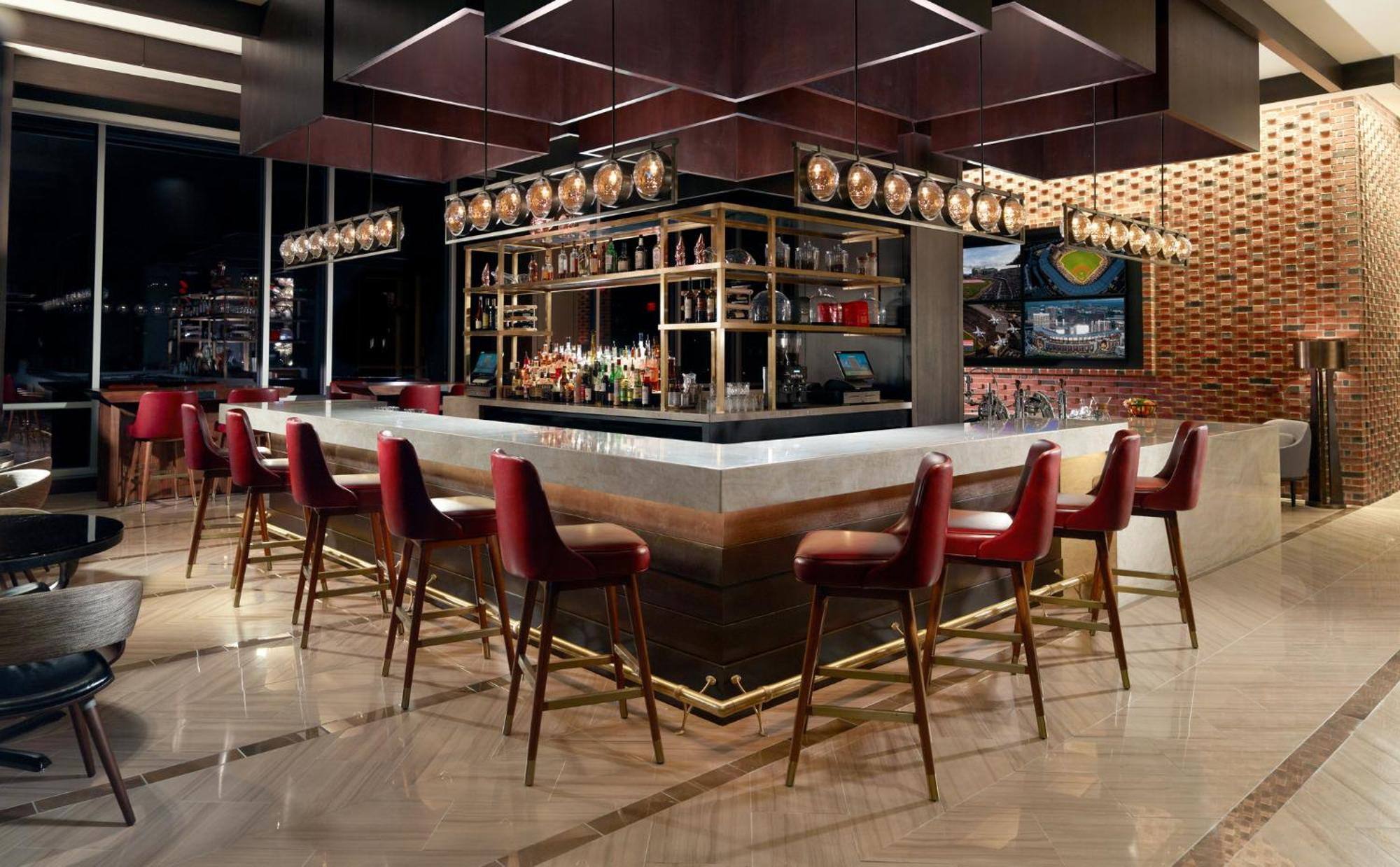 Modern hotel bar with a square marble counter, red barstools, shelves of liquor bottles, and contemporary lighting in a stylish indoor setting.