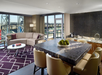 Modern hotel suite with a curved sofa, geometric rug, marble dining table with chairs, kitchenette, and large windows overlooking a stadium.
