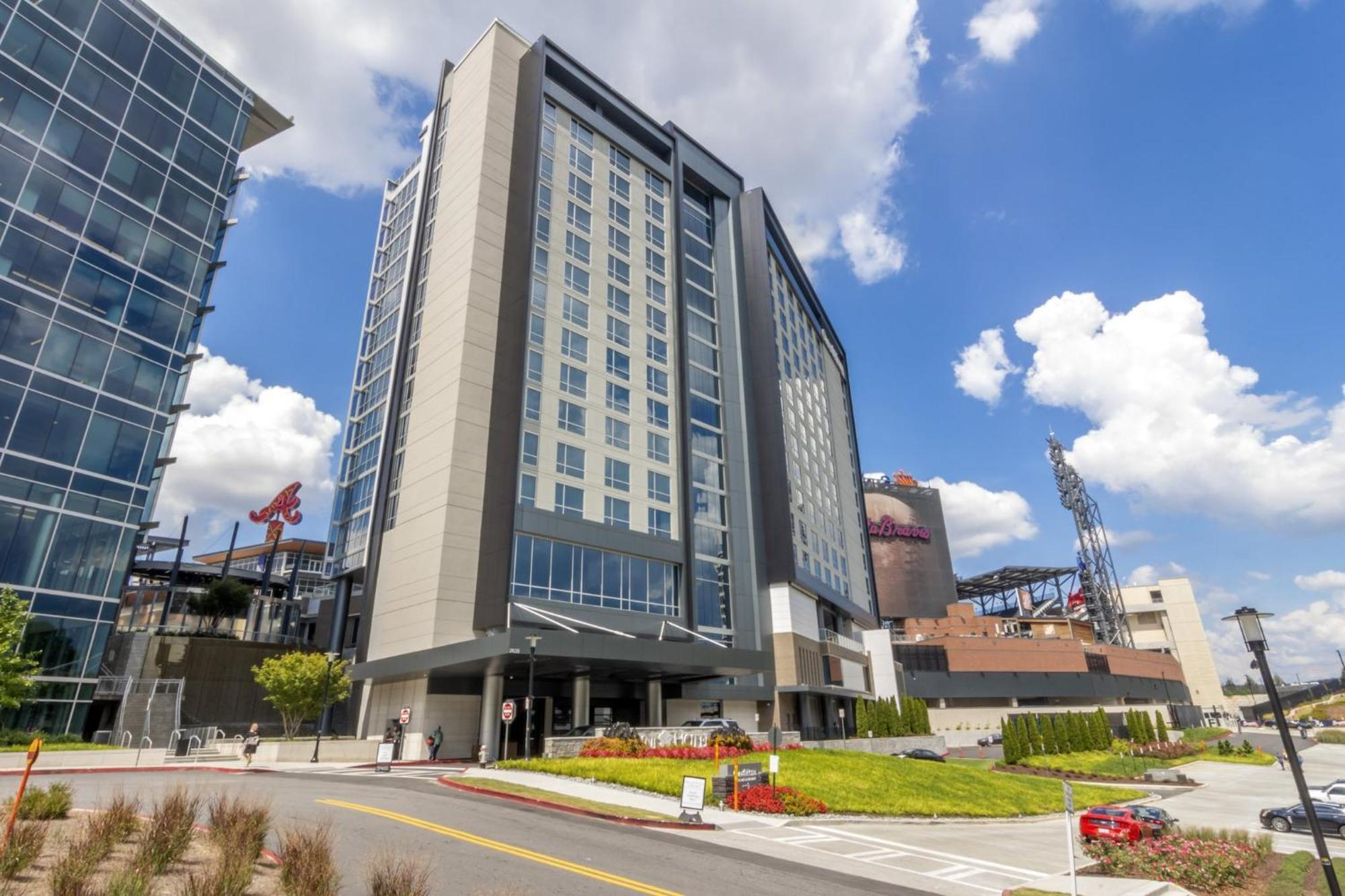 Tall modern hotel building with large windows, located near a stadium with an Atlanta Braves "A" logo, under a blue sky with scattered clouds.