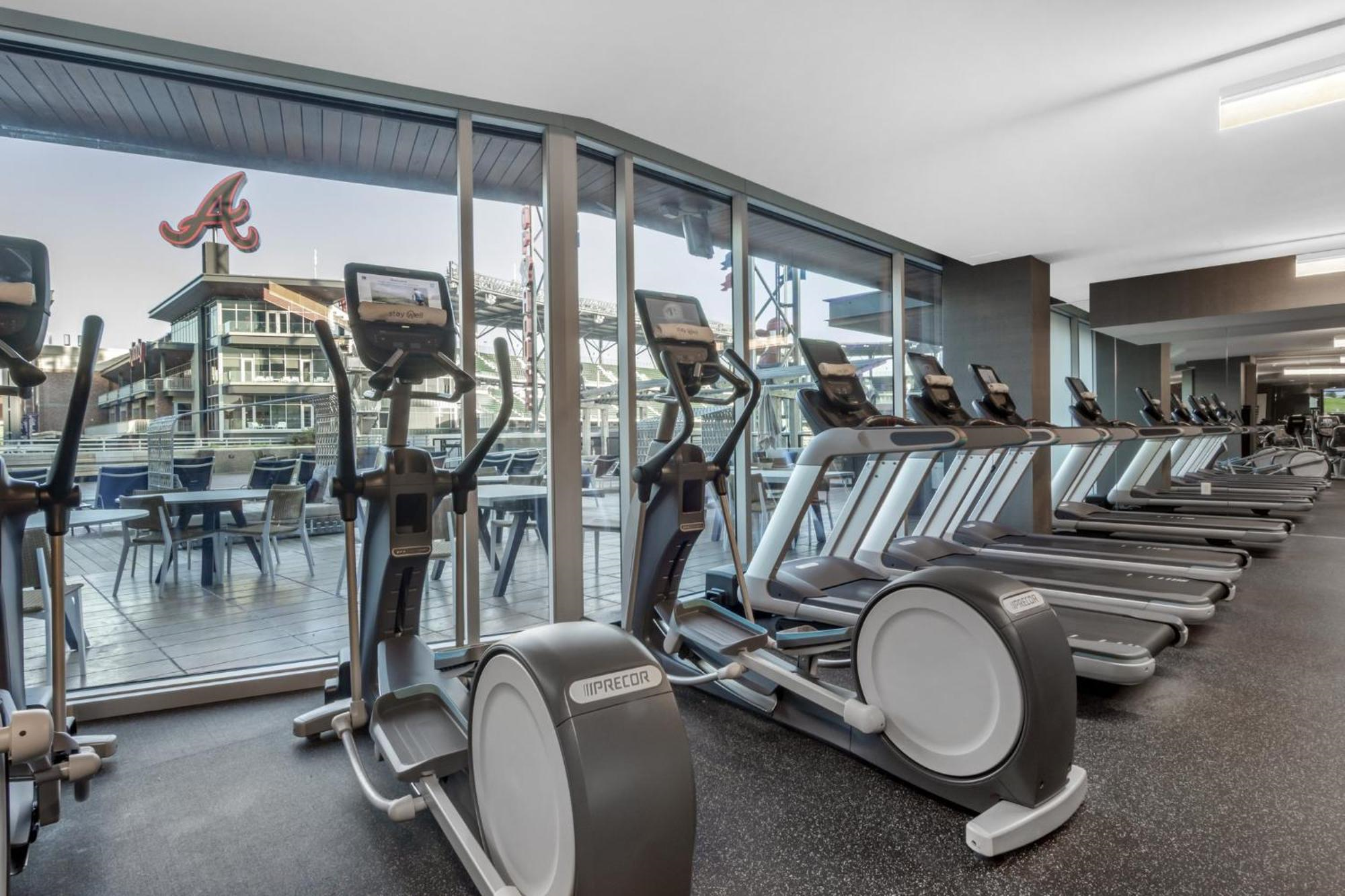 Row of elliptical machines and treadmills in a modern gym with large windows overlooking an outdoor patio and buildings with Braves signage.