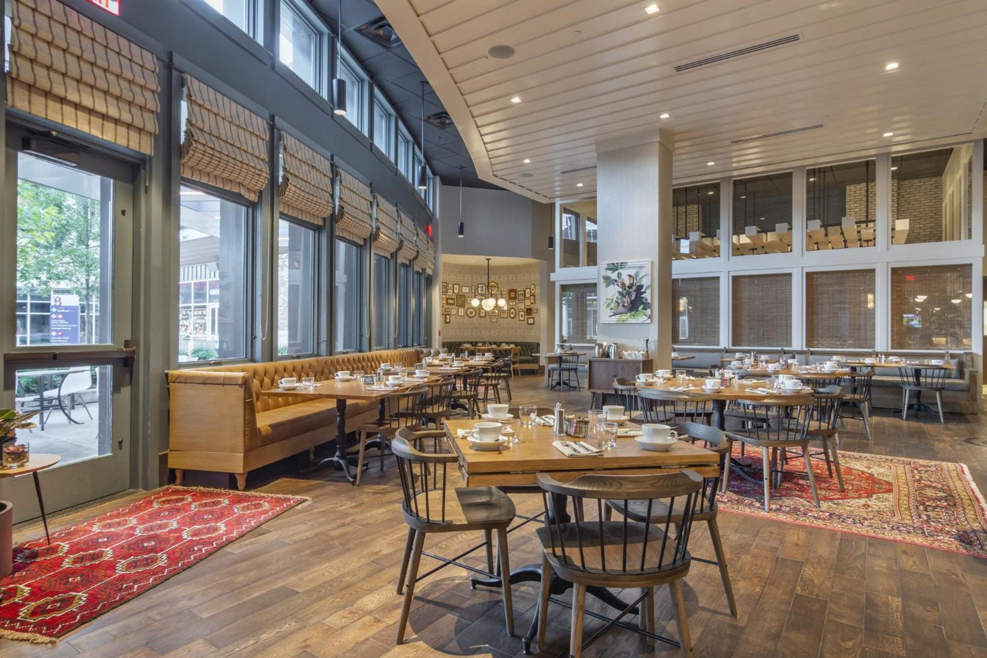Spacious restaurant dining area with wooden tables and chairs, large windows, a long bench seat, and decorative rugs on a wooden floor.