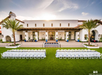 Outdoor event setup with white tables and chairs on green lawn, facing a large white building with palm trees and a fountain, under a partly cloudy sky.