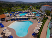 Aerial view of a resort featuring multiple outdoor swimming pools surrounded by blue loungers and white umbrellas, with a lush landscape and hills in the background.