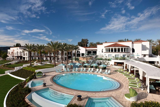 Luxurious resort with a circular pool, surrounded by palm trees and sun loungers, against a backdrop of clear blue sky. White buildings include balconies and large windows.