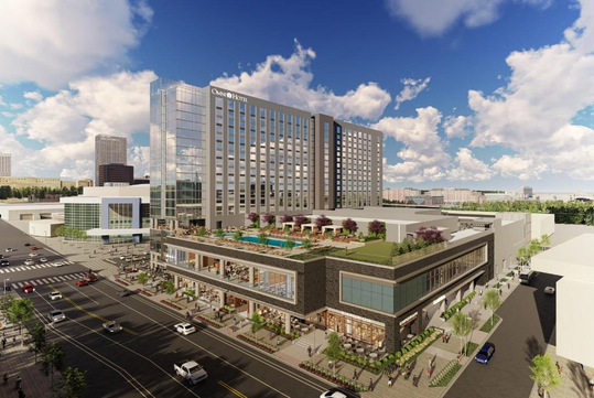 Aerial view of a multi-story hotel with adjacent buildings, a rooftop pool, and terrace. Streets with cars and pedestrians surround the complex under a partly cloudy sky.