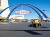 View of the back of a yellow GoCar driving down the road with a pink Las Vegas sign hanging over the road on a sunny day.