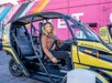 A woman in a brown leather jacket smiling and sitting in a yellow doorless GoCar with a pink wall behind her in Las Vegas.