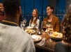 A group of friends gathered around a table enjoying dinner and drinks at the One World Observatory with the windows behind them showing NYC.