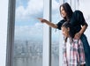 A mother and daughter standing close to a window at the One World Observatory pointing and looking down at New York City.