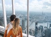 A man and woman holding each other and looking out of the windows of the One World Observatory down on NYC on a sunny day.