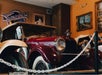 A vintage red and white car is displayed in a museum setting, surrounded by paintings and a chain barrier.