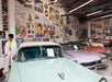 A man in retro clothing stands by classic cars in a museum filled with vintage posters and memorabilia on the walls.