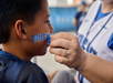 A person applies blue face paint spelling "PRIDE" on a boy's cheek at what appears to be a sports event.