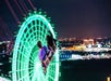 Two people on a swing ride at night, with a brightly lit green Ferris wheel and city lights in the background.