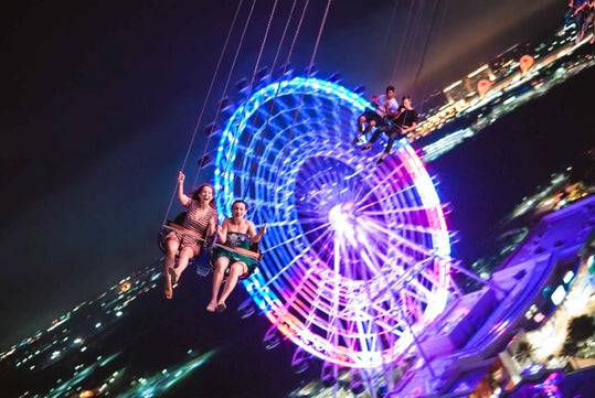 People enjoy a swing ride at night with a brightly lit Ferris wheel in the background. The city lights create a vibrant atmosphere.