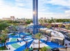 Aerial view of an amusement park ride surrounded by people and palm trees, with city buildings in the distance under a blue sky.