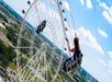 A person rides a swing carousel next to a large Ferris wheel with a bright blue sky and scattered clouds in the background.