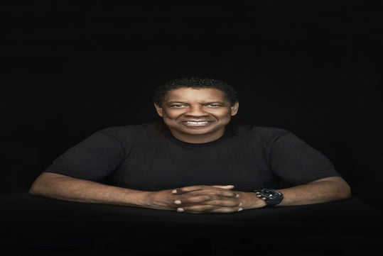 Man in a black shirt smiling and sitting with hands clasped against a dark background.