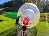 Two people are outdoors; one sits on the grass in front of a large clear OGO zorb ball while the other stands nearby, reaching out a hand.