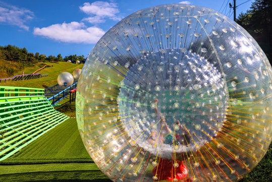 A large, transparent inflatable ball for zorbing sits on a grassy outdoor track under a blue sky with scattered clouds.