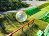 A person rolls downhill inside a large transparent inflatable zorb ball at an outdoor adventure park with green ramps and grassy slopes.