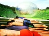 Several people lay face down in a row on a green outdoor ramp as a large inflatable zorb ball rolls toward them from the background.