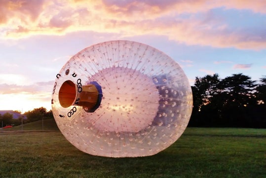 A large transparent inflatable zorb ball sits on a grassy field during sunset, with trees and a colorful sky in the background.