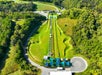 Aerial view of a green hill with two parallel grass-covered summer tubing slides, conveyor belts, canopies at the bottom, and surrounding trees and parking area.