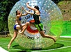 Two people in swimsuits jump and high-five in front of a large inflatable zorb ball on a grass field under sunny weather.