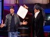 A person in a cap laughs while another blindfolded person holds up a drawing of a vase on a whiteboard.