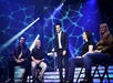 A man in a suit stands on a stage with four seated people under blue lighting effects.