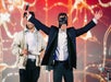 Two people on stage. The person on the right wears a black mask and raises their arms, holding a microphone. The other person stands beside them, holding a cloth. Colorful backdrop behind.