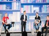 Four people are seated on a stage with a backdrop resembling a library. One man in the center is standing and speaking, while the others sit comfortably, two wearing red shirts.