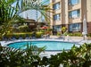 Outdoor swimming pool surrounded by lounge chairs and tropical plants next to a multistory hotel building under a sunny sky.