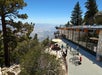 View looking down at people sitting at picnic tables at The Peaks patio on a sunny day with trees around them and mountains in the distance.