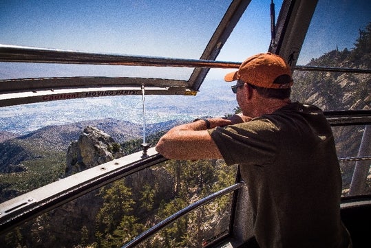 A man in an orange baseball cap inside the tramcar leaning on the railing of an open window looking at the mountains at Palm Springs Aerial Tramway.