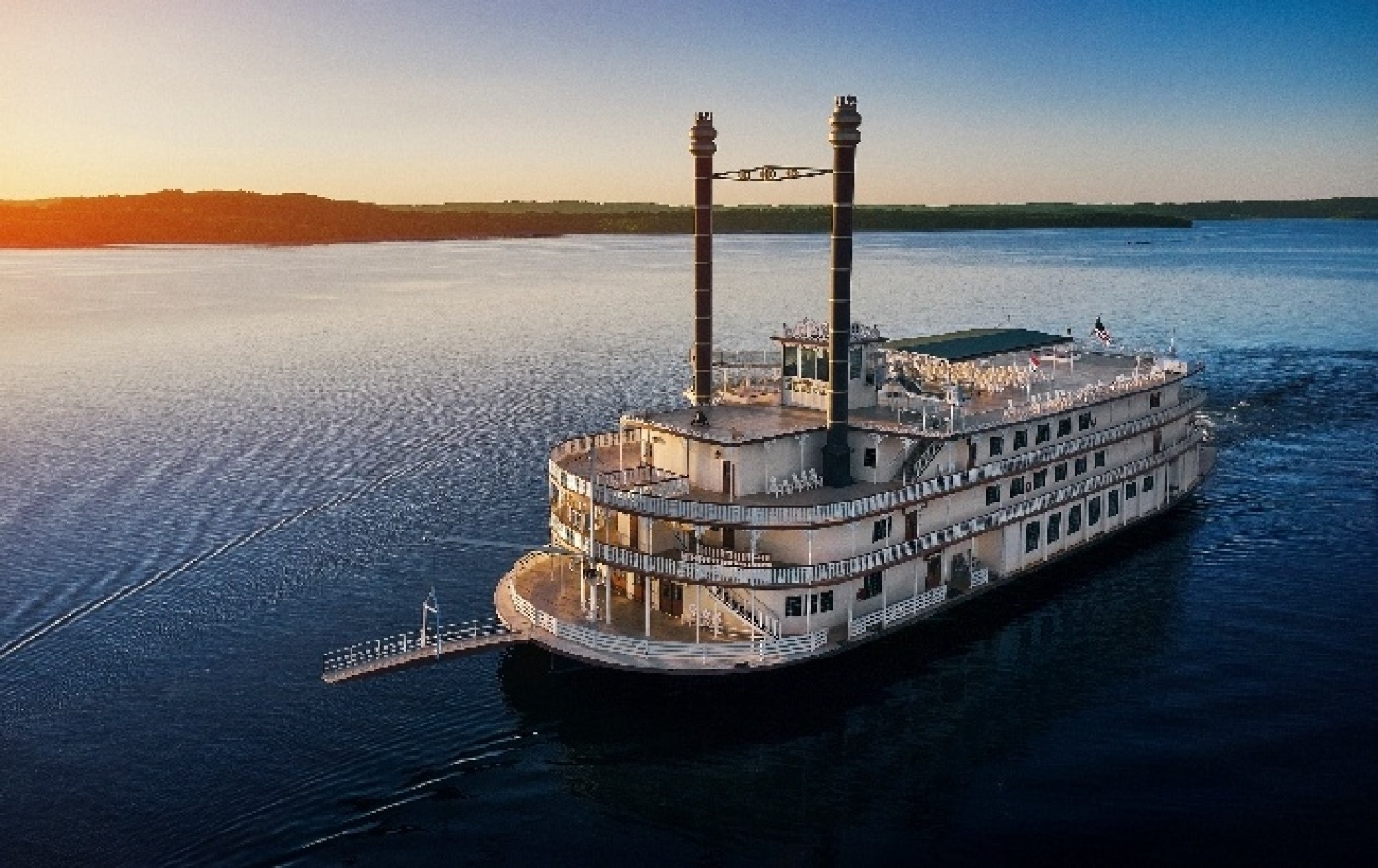A white paddle steamer with two black smokestacks sails on a calm, expansive body of water under a clear sky at sunset.