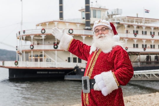 A man dressed as Santa Claus stands by a river, gesturing towards a large, decorated paddlewheel steamboat.