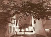 Sepia-toned photo of a narrow alley with old buildings on both sides, tree branches overhead, and streetlights illuminating the path at night.