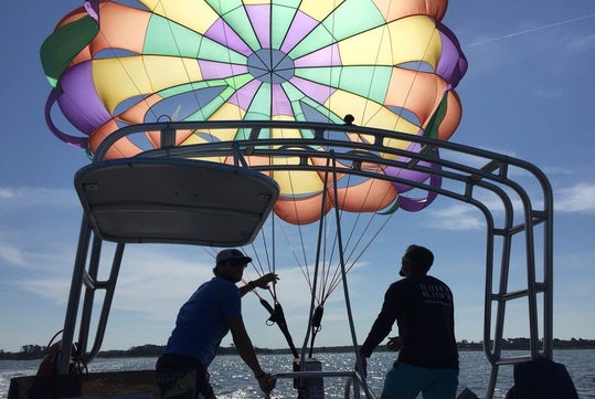 Two people on a boat guide a bright, multicolored parasail in the air, with one person pointing and the other holding the controls. The sky is clear, and the sea is calm.