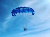 Two people parasailing under a blue parachute against a clear sky.