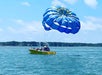 A yellow boat labeled "ISLAND HEAD" on the water with people aboard and a blue and yellow parachute attached to the boat, partially inflated in the air against a background of trees and a blue sky.