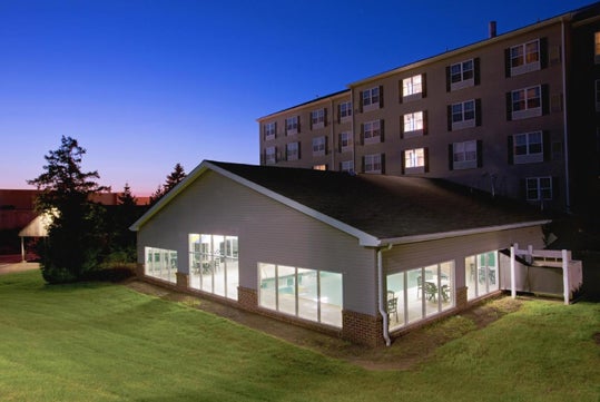 A brightly lit building with large windows sits next to a multi-story apartment complex at dusk, surrounded by grass and trees.