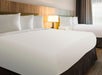 Hotel room with two neatly made beds, white linens, wooden headboard, and a lit bedside lamp. Curtain-covered window on the right.