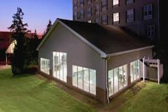 A large building with glass walls and a gable roof is surrounded by grass, adjacent to a tall residential or office building, during dusk.