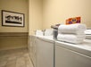 Laundry room with a row of white machines, folded towels, a box of Tide detergent, tiled floor, and framed wall art of trees.